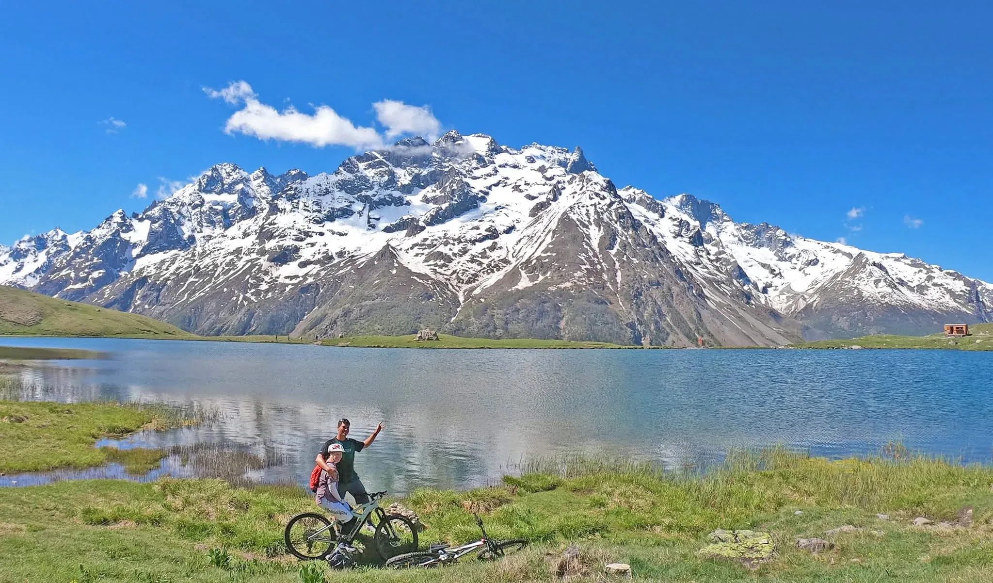 Two mountain bikers beside their bikes at an Alpine lake beneath snow-covered peaks