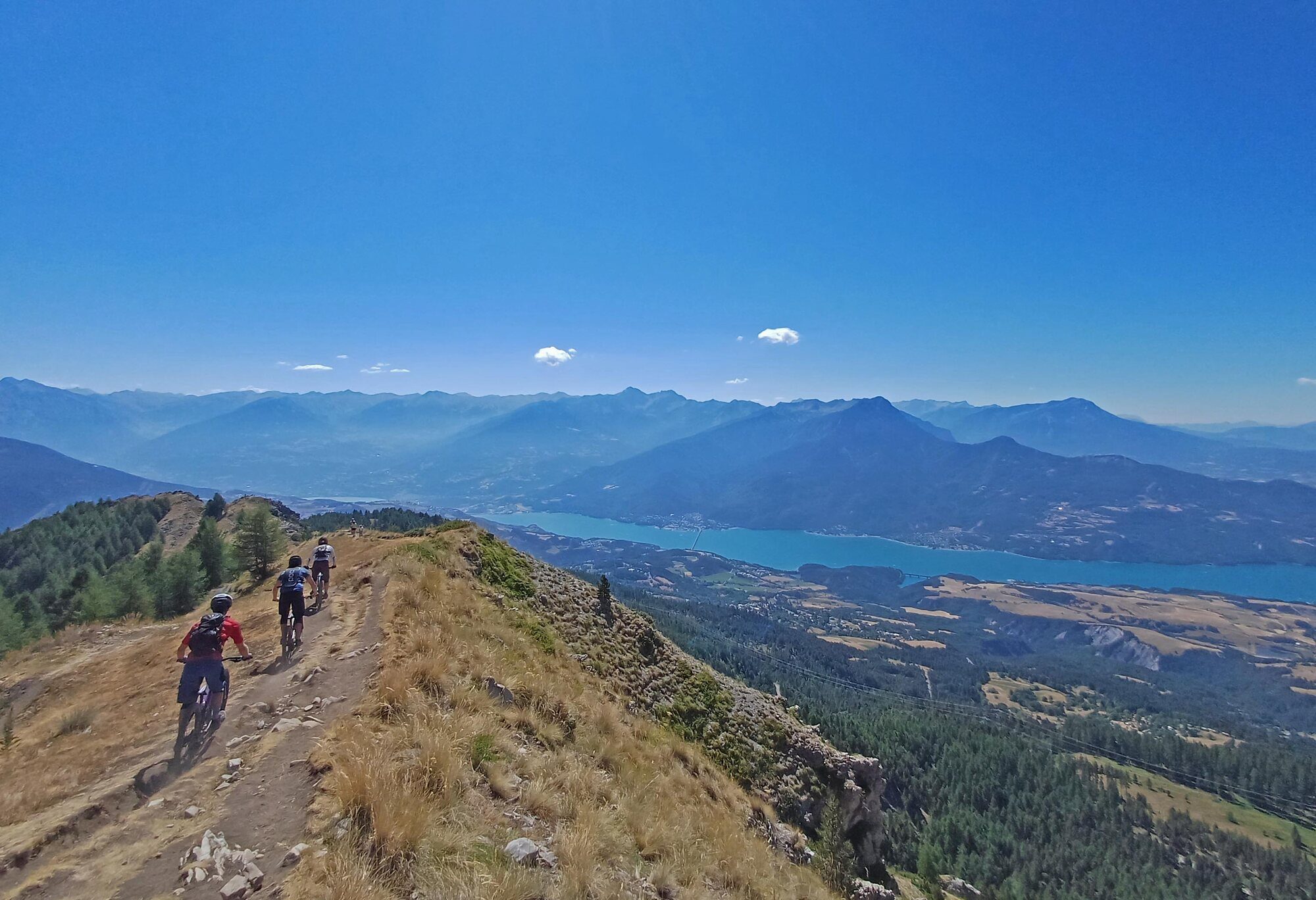 Three mountain bikers riding a rocky trail along a grassy Alpine ridge above a blue lake