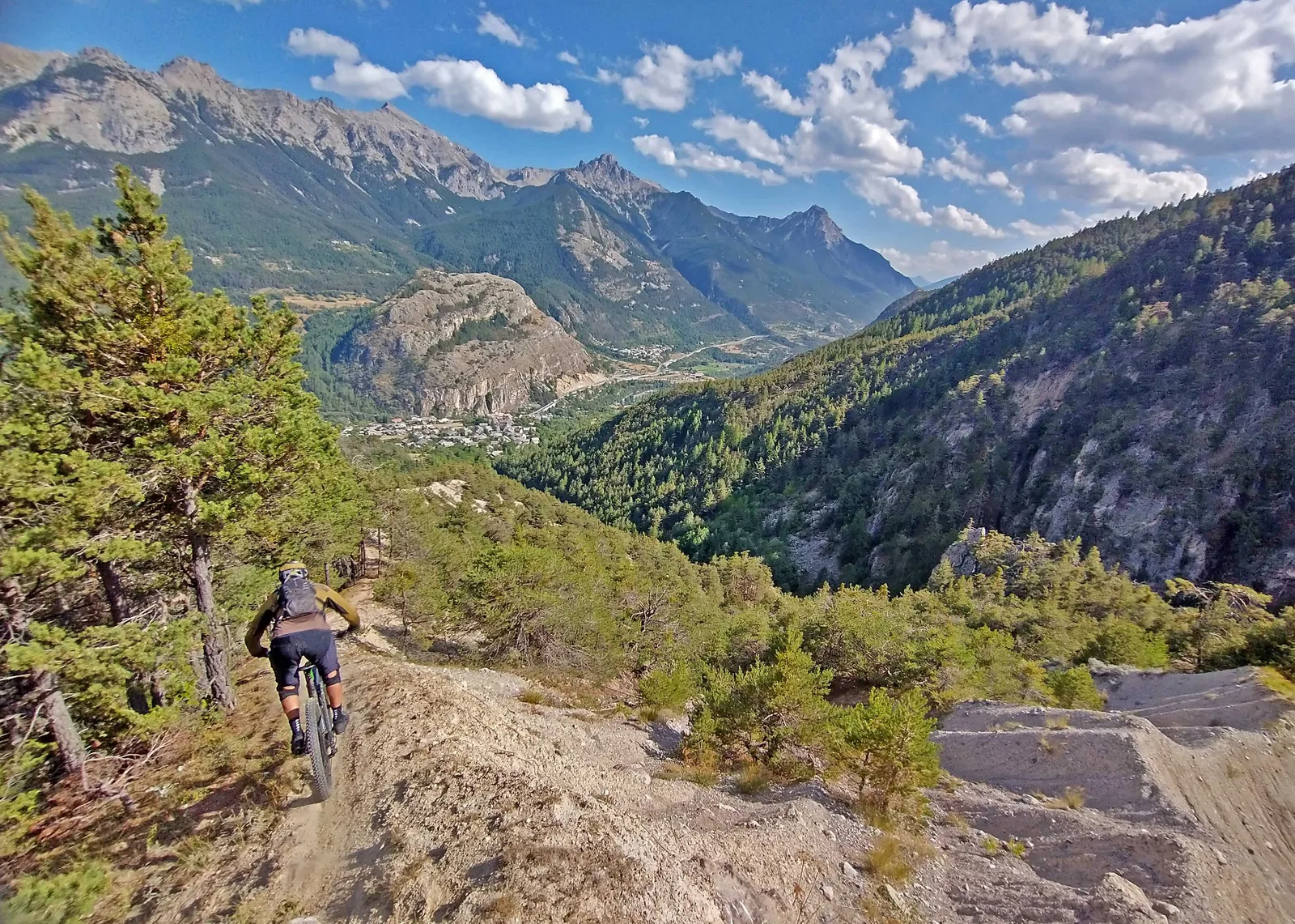 Mountain biker descending a rocky singletrack toward a broad Alpine valley