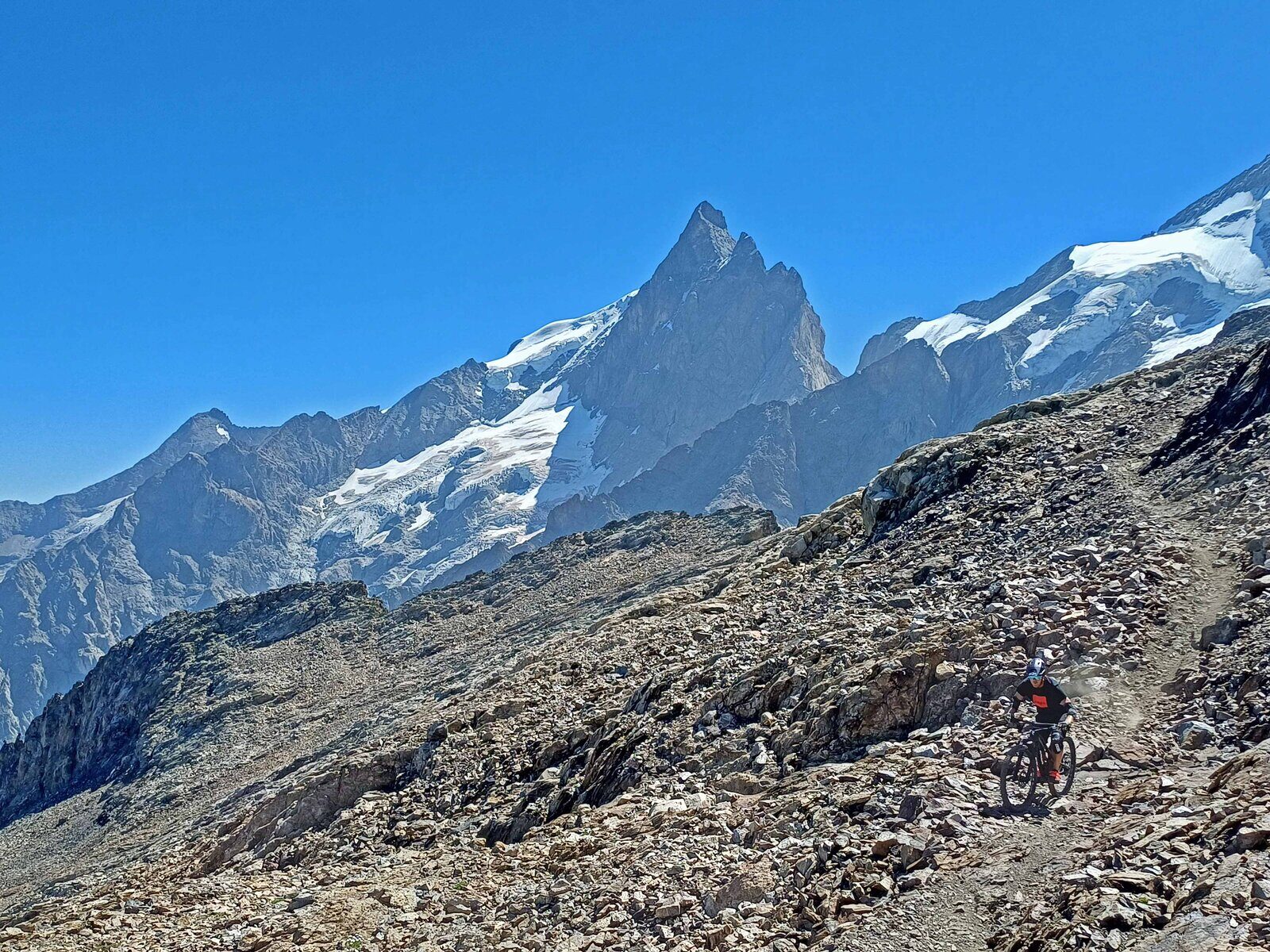 Mountain bikers riding a rocky high-Alpine trail beneath snow-capped peaks